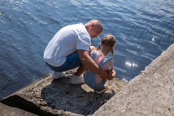 A father is crouched by his daughter at water's edge, conversation shared. Suggests guidance, care, and family bonding time, and health insurance when in nature and water, safety. High quality photo