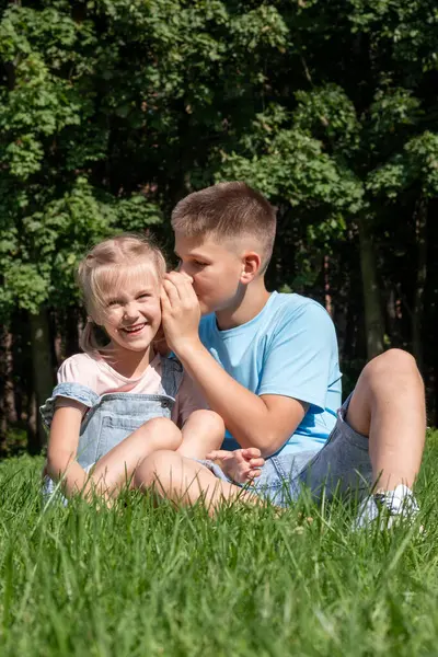 Young boy whispers to a smiling girl in a sunlit park, childhood camaraderie and trust. bond of siblings, for National Siblings Day promotions or narratives about youth friendships. High quality photo