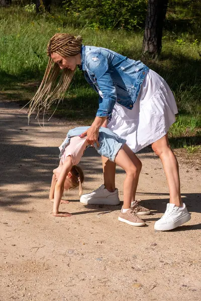 A mother supports her daughters playful upside-down bend on a forest path, sharing a moment of laughter and connection, bonding time in the nature. High quality photo