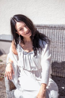 Neşeli bir gülüşü olan bir kadın beyaz, akıcı bir elbise ve hırka içinde rahatça uzanır, şık aksesuarlar ve rahat bir tavırla görünüşü tamamlanır. Yüksek kalite fotoğraf