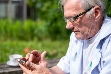 Yansıtıcı yaşlı erkek akıllı telefonuna göz atıyor, yeşil bir parkın dinginliğini dijital çağların nabzı ile birleştiriyor. Yüksek kalite fotoğraf