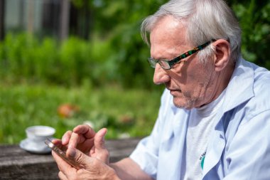 Gözlüklü gri saçlı adam dışarıda oturan mobil cihazıyla etkileşime giriyor ve nesiller boyu teknolojiyle aralarındaki köprüyü resmediyor. Yüksek kalite fotoğraf