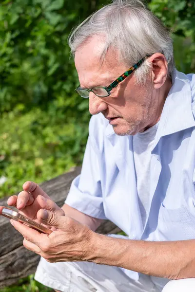 Yaşlı bir adam güneşli, yeşil bir parkta desenli gözlükleriyle akıllı telefonuna dikkatle odaklanır. Görüntü modern emeklilikte teknolojinin temalarını çağrıştırıyor. Yüksek kalite fotoğraf