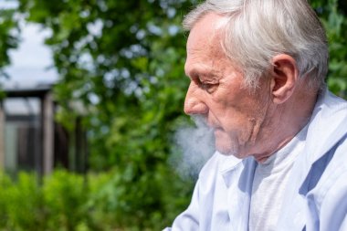 Dışarıda sigara içen düşünceli yaşlı bir adamın profili. Arka planda yeşil yapraklar var. Yalnızlığı yansıtıyor. Yüksek kalite fotoğraf