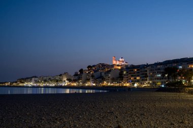 Sakin bir sahilde alacakaranlık, şehrin ışıkları ve arka planda aydınlık bir katedral, huzurlu bir akşam sahnesi. Yüksek kalite fotoğraf
