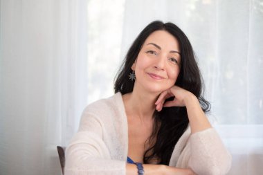 Kendine güvenen orta yaşlı bir kadın, uzun siyah saçlı, rahat beyaz bir kazakla gülümseyen, aydınlık bir odada, sakinlik kavramını ifade eden