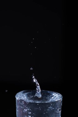 Temiz bir bardaktan su sıçrıyor, dinamik damlacıklar yaratıyor ve bu damlalar karanlık bir zemin üzerinde yükselip düşüyor, hareketli suyun güzelliğini gösteriyor..