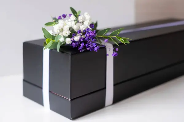 Güzel paketlenmiş siyah bir hediye kutusu bir yüzeyde duruyor, mor ve beyaz tonlarında taze çiçeklerle süslenmiş, her kutlama için sofistike bir dokunuş sunuyor..