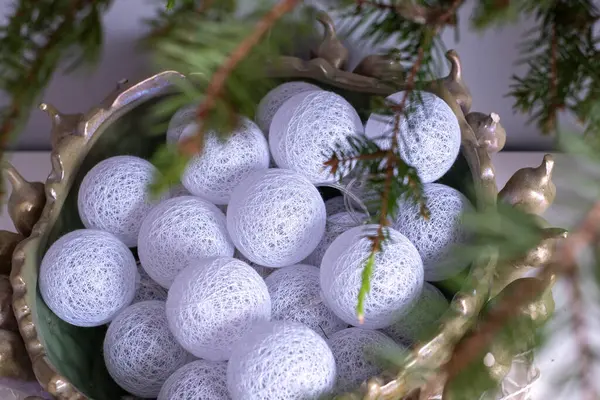 Zarif beyaz dekoratif küreler, kısmen yeşil dallar tarafından gizlenmiş yeşil bir kaseyi doldurur ve kış dekorunda sıcak bir şenlik ortamı yaratır..