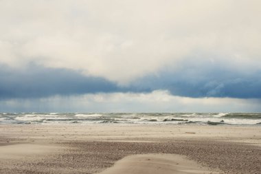 Yumuşak dalgalar bulutlarla dolu kasvetli bir gökyüzünün altındaki kumlu bir sahile çarpıyor. Atmosfer sakin ama dramatik. Huzurlu bir yürüyüş için mükemmel..