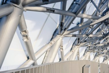 Metal kirişlerin iskeleti gelişmiş mühendislik ve tasarım sergiliyor, kentsel bir alanın üstündeki parlak ve berrak gökyüzüne karşı çarpıcı bir görsel element oluşturuyor..