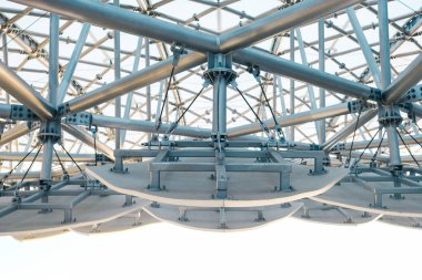 Geniş bir mimari tepe örtüsünün altında, parlak bir arkaplan karşısında modern mühendislik ve tasarım sergilenen karmaşık bir metal destek ağı bulunur..