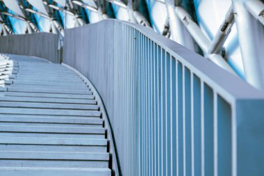 Çağdaş bir merdiven, güneşli bir günde parlak mavi ve metalik yapılardan oluşan bir zemin üzerine kurulmuş pürüzsüz, eğri bir tırabzan ve doğrusal adımlar içerir..