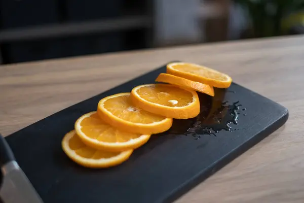 Yeni dilimlenmiş turuncu segmentlerin canlı bir görüntüsü parlak siyah bir kesim tahtasına yerleştirilmiştir. Doğal ışık dolu modern bir mutfak atmosferini yansıtır..