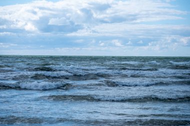 Gün ortasında sakin bir kıyı şeridinde bulutların sürüklenmesi ve sakin bir atmosfer oluşturması gibi yumuşak dalgalar kumlu sahile vuruyor..