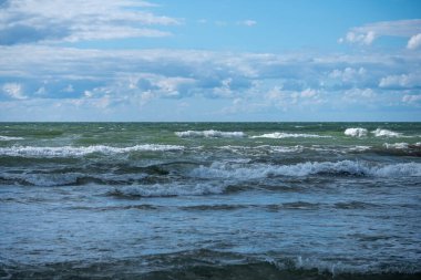 Yumuşak dalgalar kumlu kıyı şeridinde süzülüyor, huzurlu bir sahil manzarasında, parçalı bulutlu bir gökyüzü altında huzur verici bir ses yaratıyor..