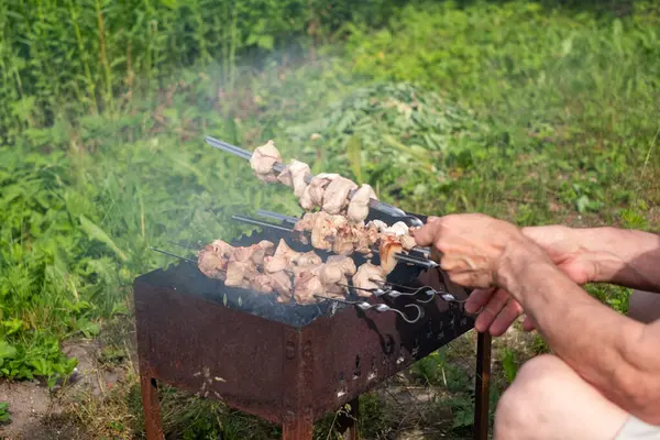 Et şişleri paslı bir barbeküde kızgın kömürlerin üzerinde ızgara yapıyor. Bir insan yeşil bir bahçede şişler ile ilgileniyor, açık havada canlı bir günün tadını çıkarıyor..