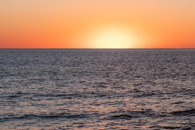 Güneş ufukta batar, turuncu ve sarı tonlarda çarpıcı bir görüntü yaratır. Sakin okyanus, sönen ışığı yansıtır ve akşam sükunetini arttırır..