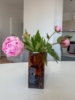 Güzel pembe şakayıklar, beyaz bir masadaki eşsiz kahverengi bir vazoda sergilenir. Çağdaş yaşam alanına yeşillikle canlı bir dokunuş eklenir.