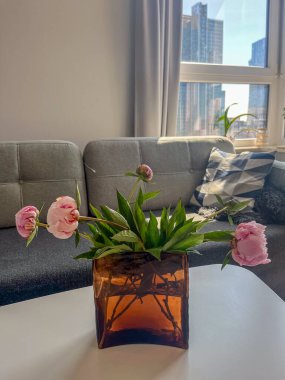 Güzel pembe şakayıklar beyaz bir masanın üzerinde kahverengi camdan bir vazoda, rahat bir oturma odası zeminine kurulmuş gri bir kanepe ve pencereden görünen şehir silüeti