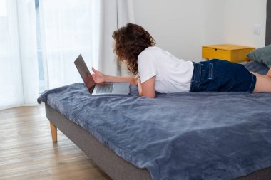 Serbest çalışan kadın dizüstü bilgisayar kullanıyor. Yatakta uzanırken, rahat bir yatak odasında, doğal ışıkla, uzaktan çalışma ve rahatlama kolaylığını gösteriyor.