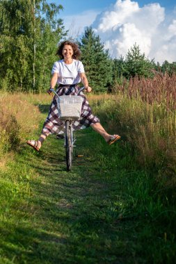 Kadın bisikletçi güneşli bir günde bisiklet sürüyor, yemyeşil ve uzun otlarla çevrili, doğadaki neşe ve refahın özünü yakalıyor.