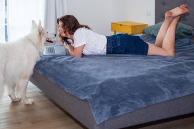 Uzaktan kumandalı kadın bir dizüstü bilgisayarla yatakta uzanıyor, sıcak ve davetkar bir yerde bir köpekle takılıyor, rahat bir iş ortamı sergiliyor.