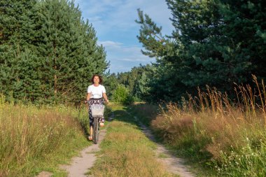 Dişi bisikletçi, canlı ağaçlar ve yabani otlarla çevrili sakin bir yolda bisiklet sürüyor. Açık hava aktivitelerinin ve refahın neşesini kucaklıyor.