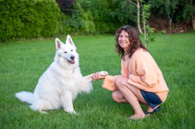 Kıvırcık saçlı bir kadın çimenlerin üzerinde diz çöküp canlı bir bahçede canlı beyaz bir köpekle meşgul oluyor. Birlikte geçirdikleri zamanda paylaştıkları bağın ve mutluluğun altını çiziyor.