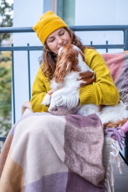 Sarı kazaklı ve şapkalı kadın balkonda oturuyor, köpeğiyle battaniyeye sarılmış, sonbahar havasının ve arkadaşlığın keyfini çıkarıyor.