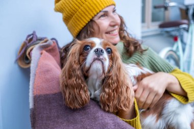 Balkonda bir kadın, köpeğiyle battaniyeye sarılmış, sonbahar sezonunun tadını çıkarıyor, sıcaklığı, arkadaşlığı ve orta yaş deneyimlerinin güzelliğini vurguluyor.