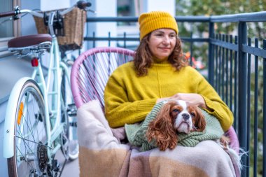 Balkonda rahat bir kadın, kalın bir kazak ve bere giyiyor, köpeğiyle vakit geçirmekten hoşlanıyor, battaniyeye sarılmış, sonbaharı sıcacık ve arkadaşça kucaklıyor.