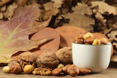 Different kinds of nuts in white cup, hazelnuts, almonds, walnuts on wooden table with autumn leaves, edible seed kernels, confectionery ingredient
