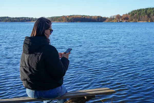 stock image Rear view of woman using mobile phone while sitting on bench at lake