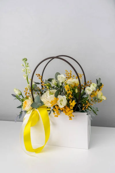 Sarı kurdeleli beyaz bir çantada güzel bir buket çiçek ve yeşillik.