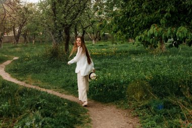 Uzun sarı saçlı, beyaz şapkalı, yeşil meyve bahçesinde yürüyen güzel bir genç kadın.