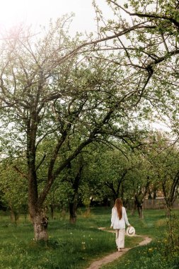 Uzun sarı saçlı, beyaz şapkalı, yeşil meyve bahçesinde yürüyen güzel bir genç kadın.