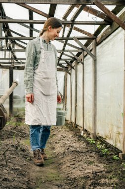 Elinde teneke kovayla serada yürüyen çekici bir kadın fidanlara bakıyor. 