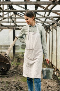 Elinde teneke kovayla serada yürüyen çekici bir kadın fidanlara bakıyor. 