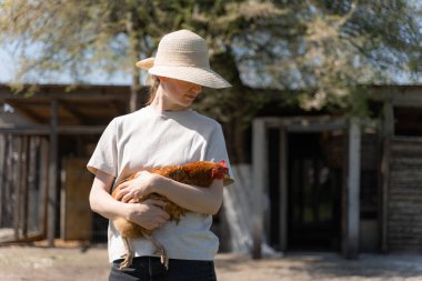 Hasır şapkalı genç kadın elinde tavuk tutuyor.