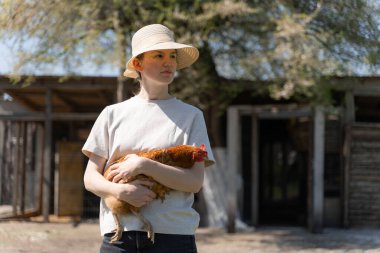 Hasır şapkalı genç kadın elinde tavuk tutuyor.