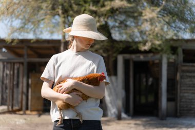 Hasır şapkalı genç kadın elinde tavuk tutuyor.