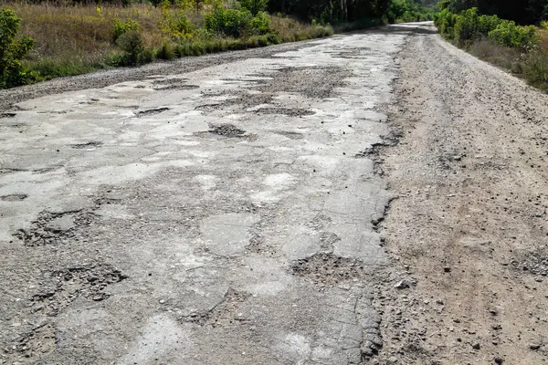 Bad road, cracked asphalt with potholes and big holes. Potholes on the road with stones on the asphalt. The asphalt surface is destroyed on the road. Bad condition of the road, needs repair.