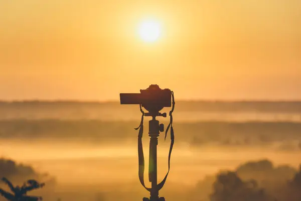 A professional photographer\'s camera on a tripod looks at the sunrise. Countryside landscape with camera on tripod, sunrise and morning fog.