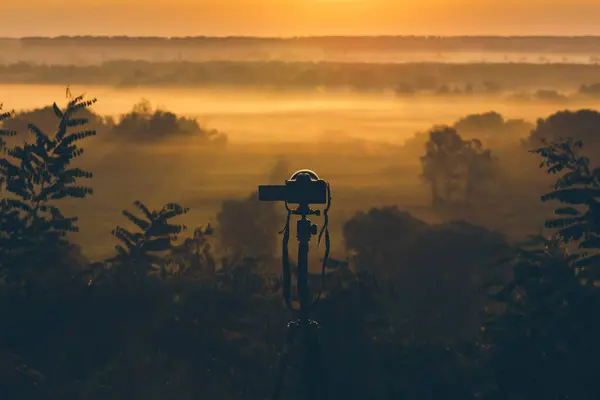 Silhouette of a camera on a tripod on a hill capturing the morning fog. Countryside landscape with a camera on a tripod, sunrise and morning fog.