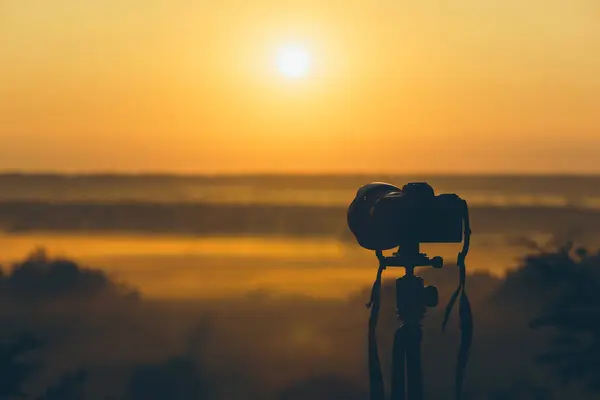 Silhouette of a camera on a tripod on a hill capturing sunrise and morning fog. Countryside landscape with camera on tripod, sunrise and morning fog.