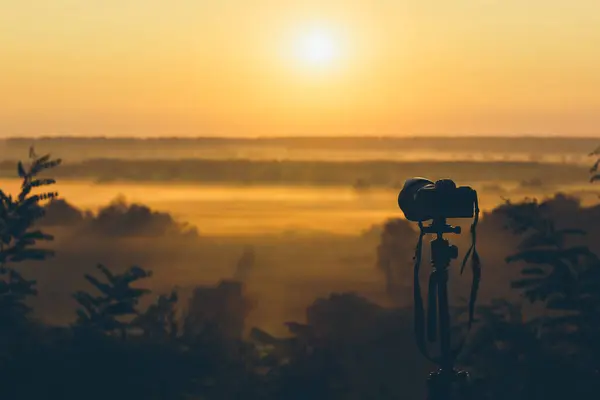 Silhouette of a camera on a tripod on a hill capturing sunrise and morning fog. Countryside landscape with camera on tripod, sunrise and morning fog.