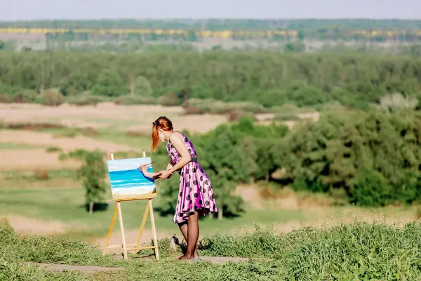 stock image A young woman is painting a landscape on a canvas while standing in nature. She is immersed in the creative process under the open sky, surrounded by a picturesque landscape