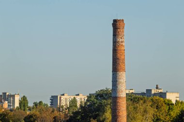 Şehrin siluetinin arka planındaki eski tuğla baca. Uzun, yıpranmış tuğla bir boru özellikle şehir binalarının ve yeşilliklerin arka planında duruyor.