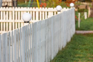Dekoratif lambalı beyaz çit. Küresel dekoratif lambalarla çevrili beyaz bir çitin yakın görüntüsü bulanık yeşil bir arka planda huzur ve banliyö hissi yaratıyor.